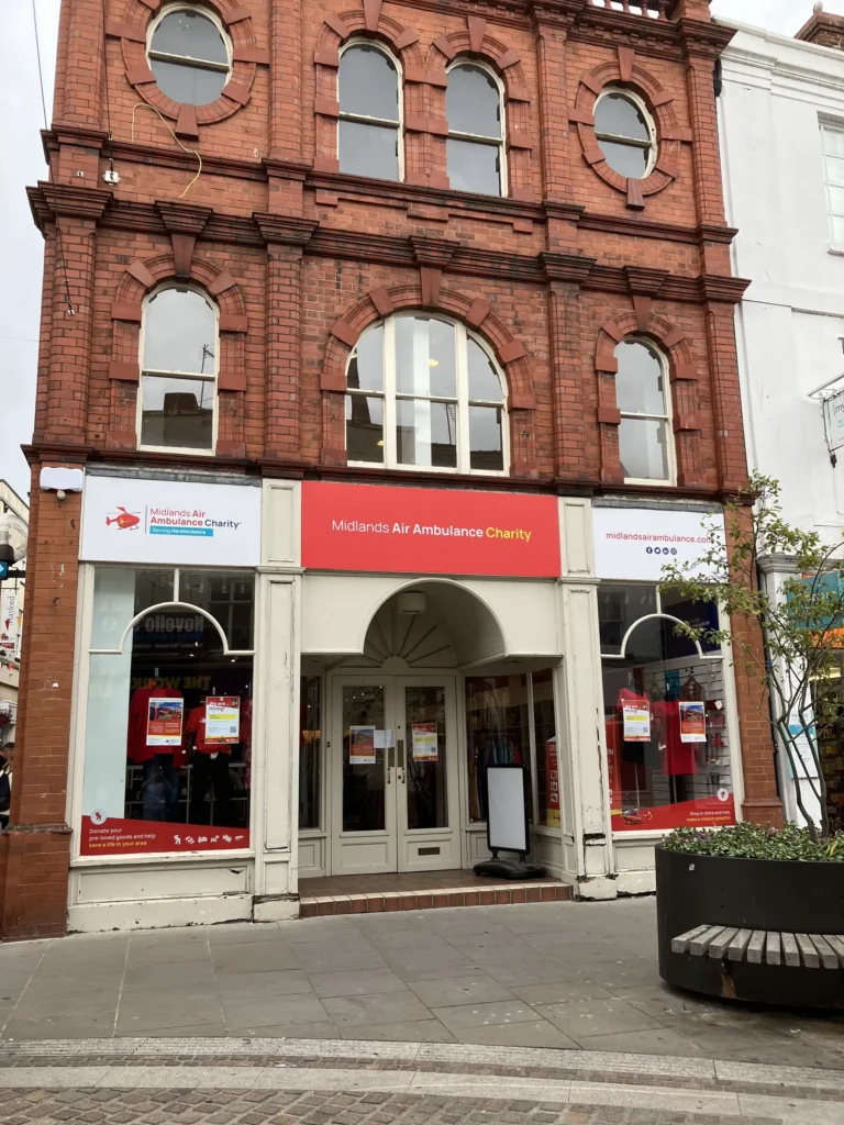 The front of Midlands Air Ambulance charity shop in Hereford, with a bright red sign and two storeys of red bricks above the shop.