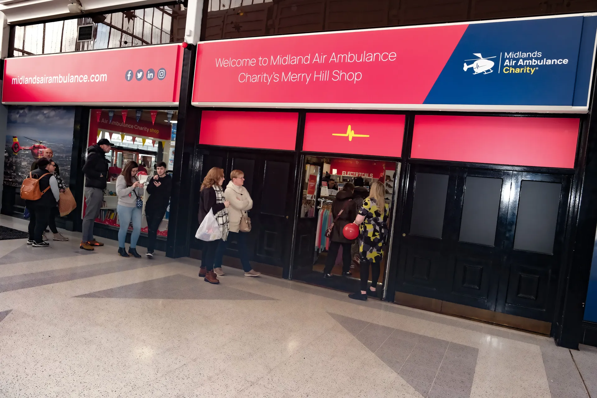The front of the Midlands Air Ambulance charity shop in Dudley, at Merry Hill shopping centre, with a bright red sign and people walking into the front door.