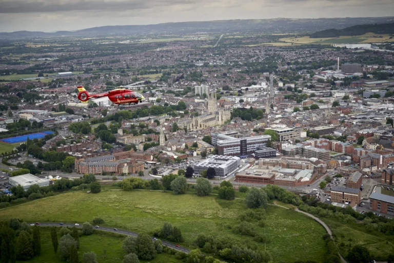 The Midlands Air Ambulance helicopter flying over Gloucestershire, with fields, housing developments and a river in the aerial view.