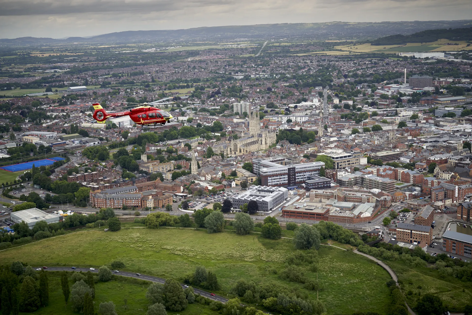 The Midlands Air Ambulance helicopter flying over Gloucestershire, with fields, housing developments and a river in the aerial view.