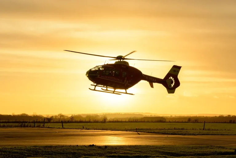 A Midlands Air Ambulance helicopter flying low over the ground with an orange sunset sky in the background.