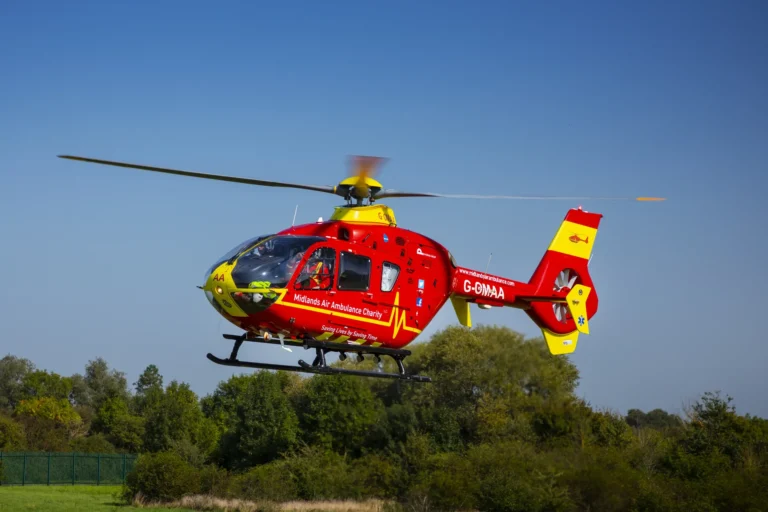 A Midlands Air Ambulance helicopter flying over a field with a blue sky and green trees in the background.