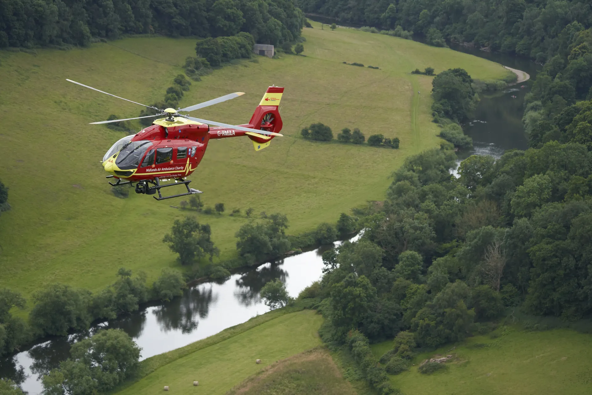 A Midlands Air Ambulance helicopter flying over Herefordshire country side, with a river running through fields.