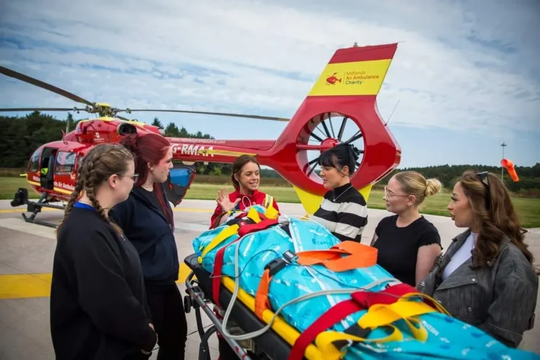 A Midlands Air Ambulance team member in red uniform leading five first aid course attendees through a first aid scenario on the helipad, with a practice patient laying on a large, yellow stretcher.