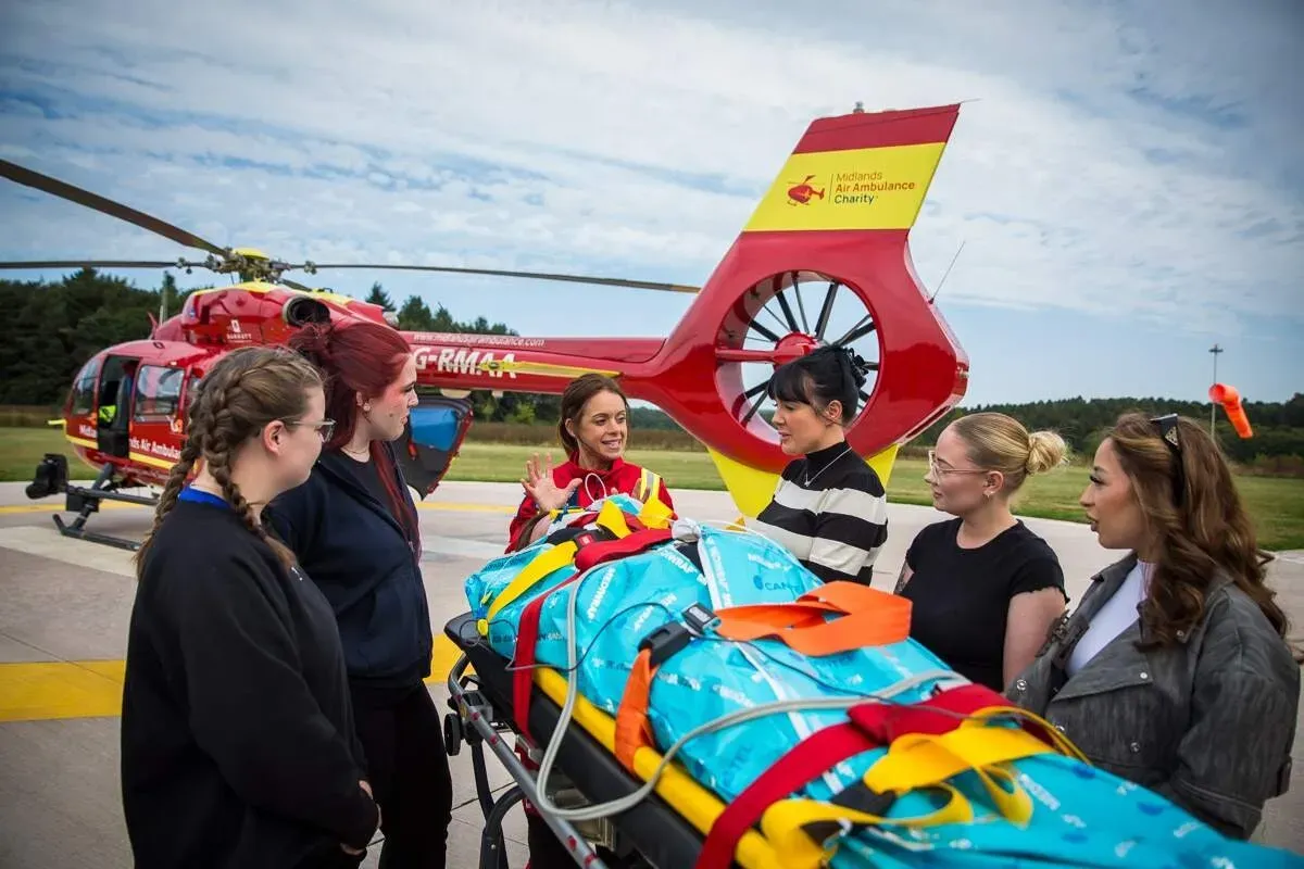 A Midlands Air Ambulance team member in red uniform leading five first aid course attendees through a first aid scenario on the helipad, with a practice patient laying on a large, yellow stretcher.