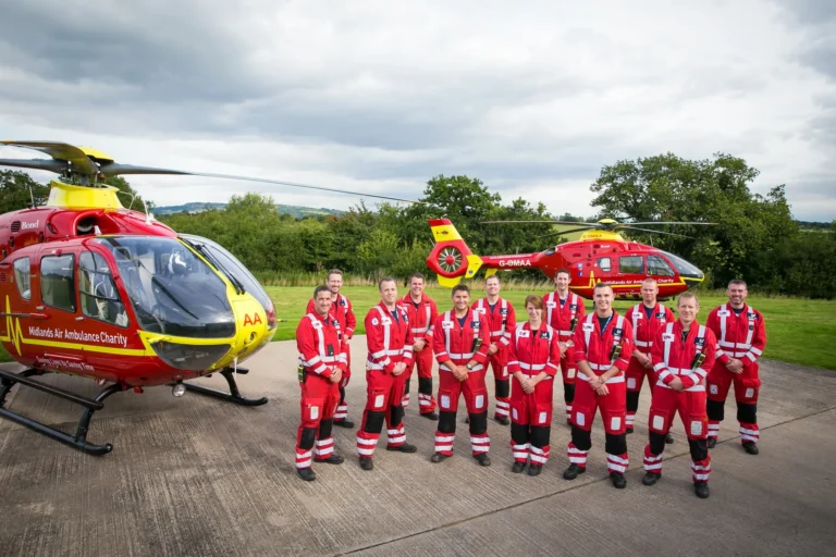 Twelve Midlands Air Ambulance Team members stood proudly on the helipad smiling in their red uniform.