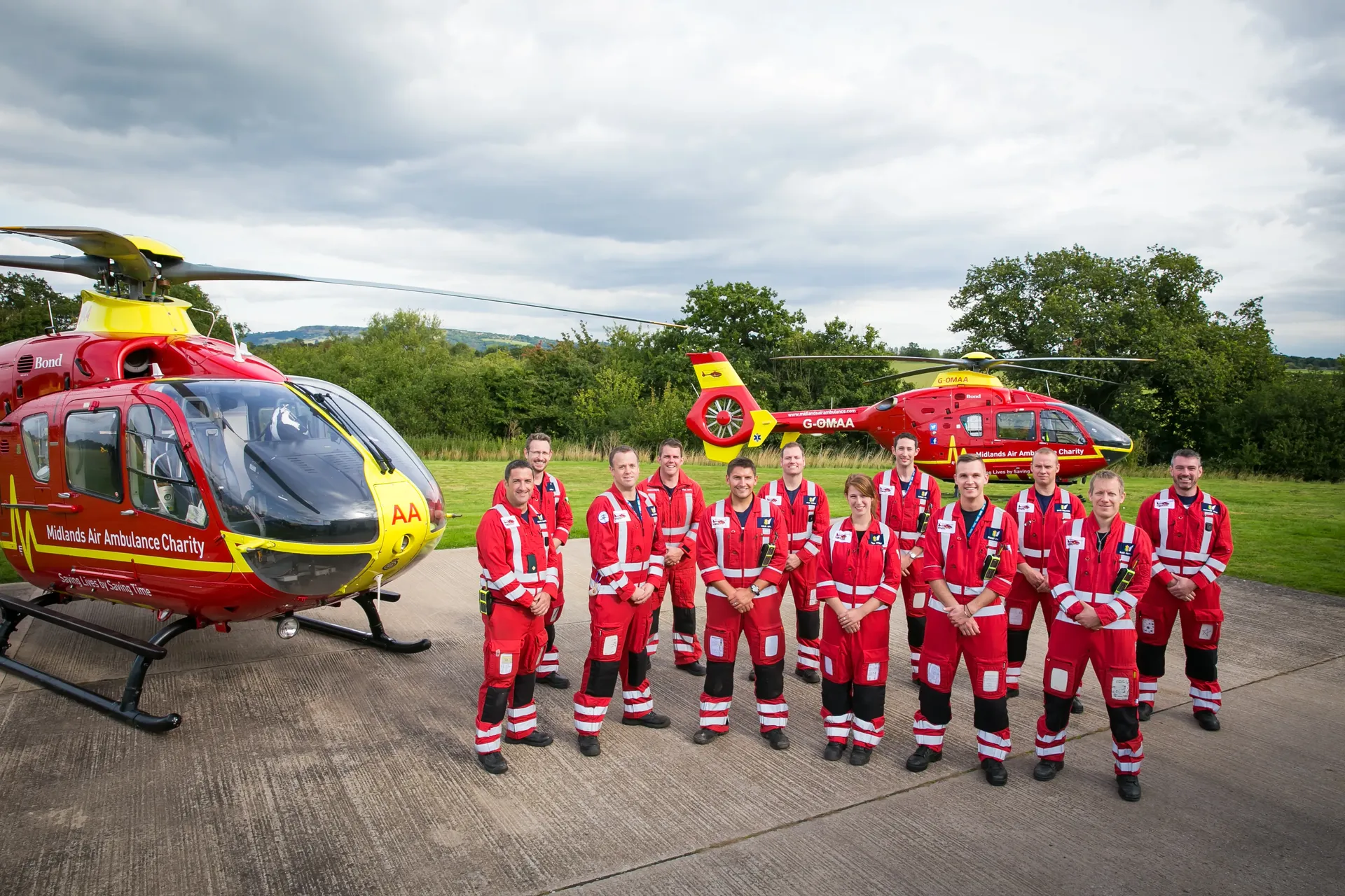 Twelve Midlands Air Ambulance Team members stood proudly on the helipad smiling in their red uniform.