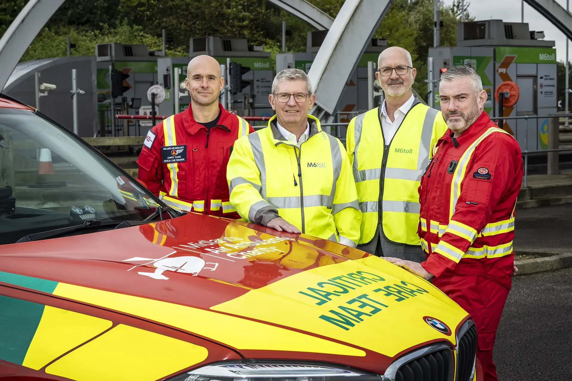 Two men in high visibility jackets from a corporate background standing in front of a Midlands Air Ambulance car with two of the Midlands Air Ambulance team dressed in red uniform standing on either side of them.