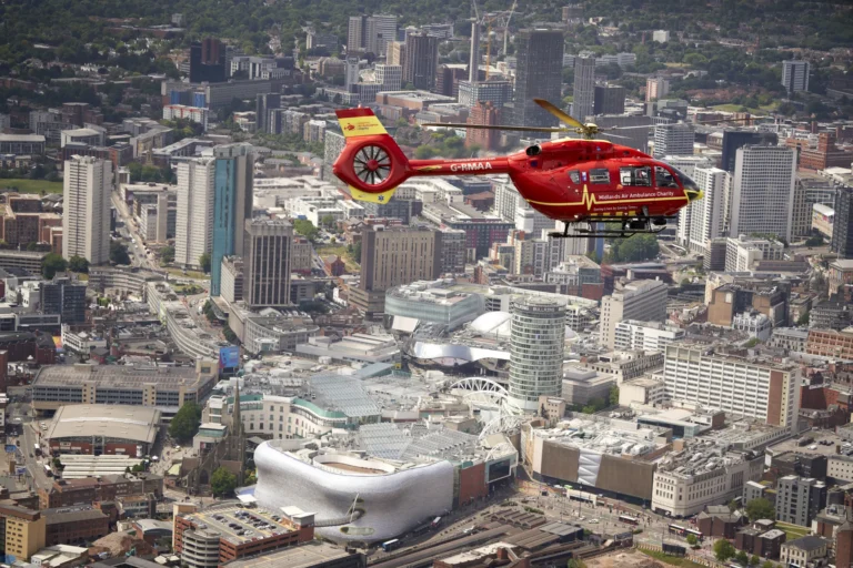 Maac West Midlands Helicopter Flying Over A City With A Large Building In The Background