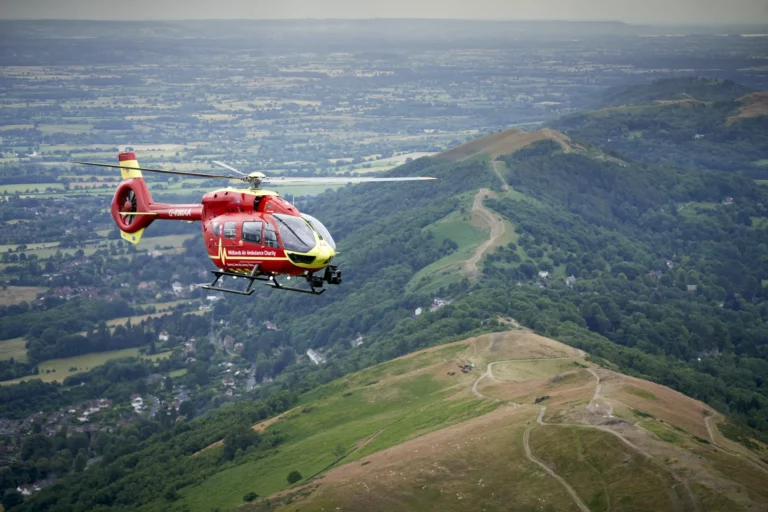 A Midlands Air Ambulance helicopter flying over Worcestershire countryside, with a path along a green peak above a valley.