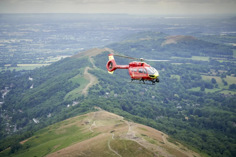 Maac Worcesteshire Helicopter Flying Over A Lush Green Hillside With A Valley In The Background