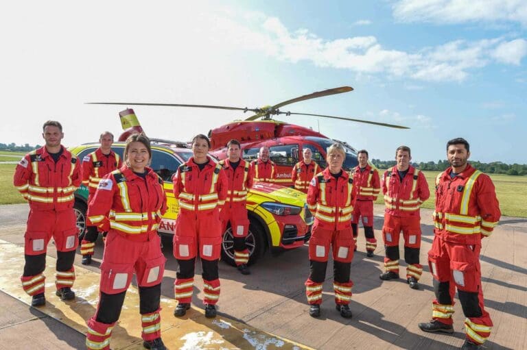 A group of ten Midlands Air Ambulance crew members stood in red and yellow uniform on the helipad with a red, Midlands car and helicopter in the background.