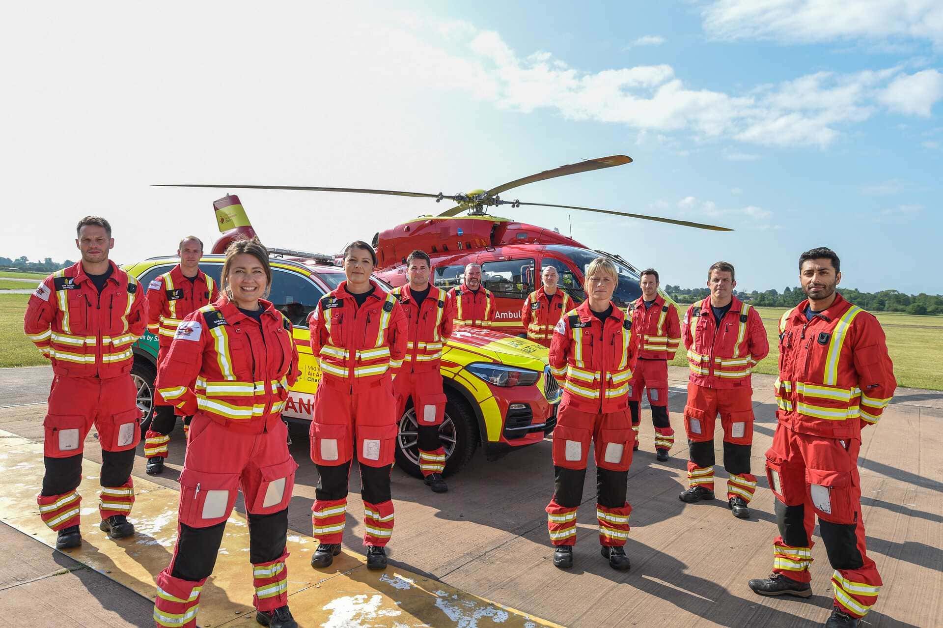 A group of ten Midlands Air Ambulance crew members stood in red and yellow uniform on the helipad with a red, Midlands car and helicopter in the background.