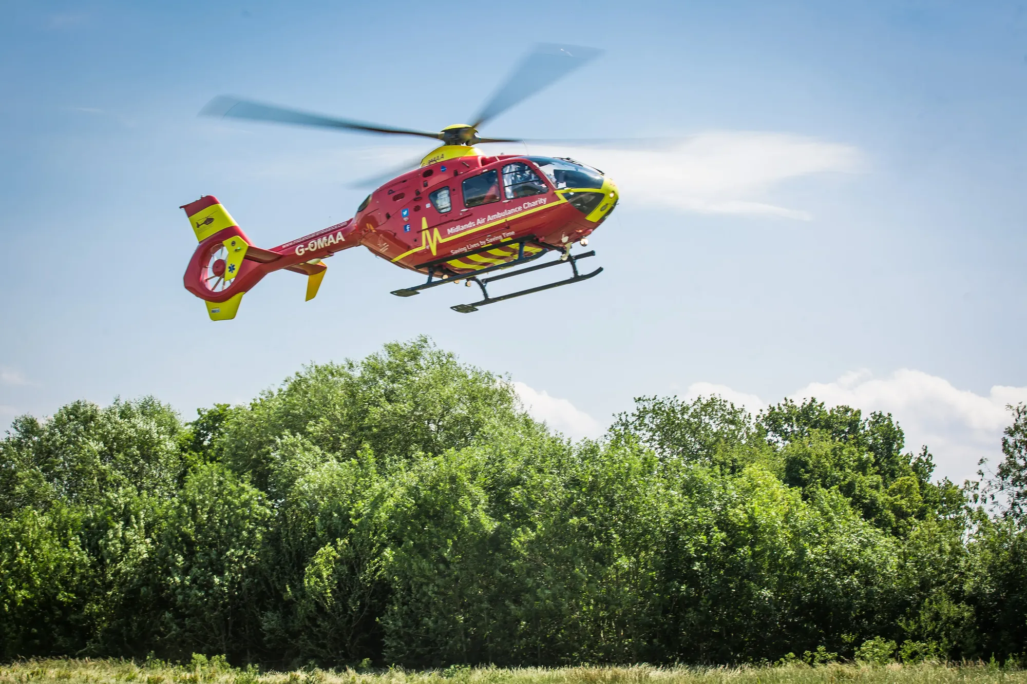 Maac Helicopter Flying Over A Field With Trees And Blue Sky