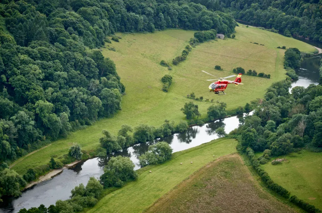 Maac Helicopter Flying Over A Lush Green Field Next To A River