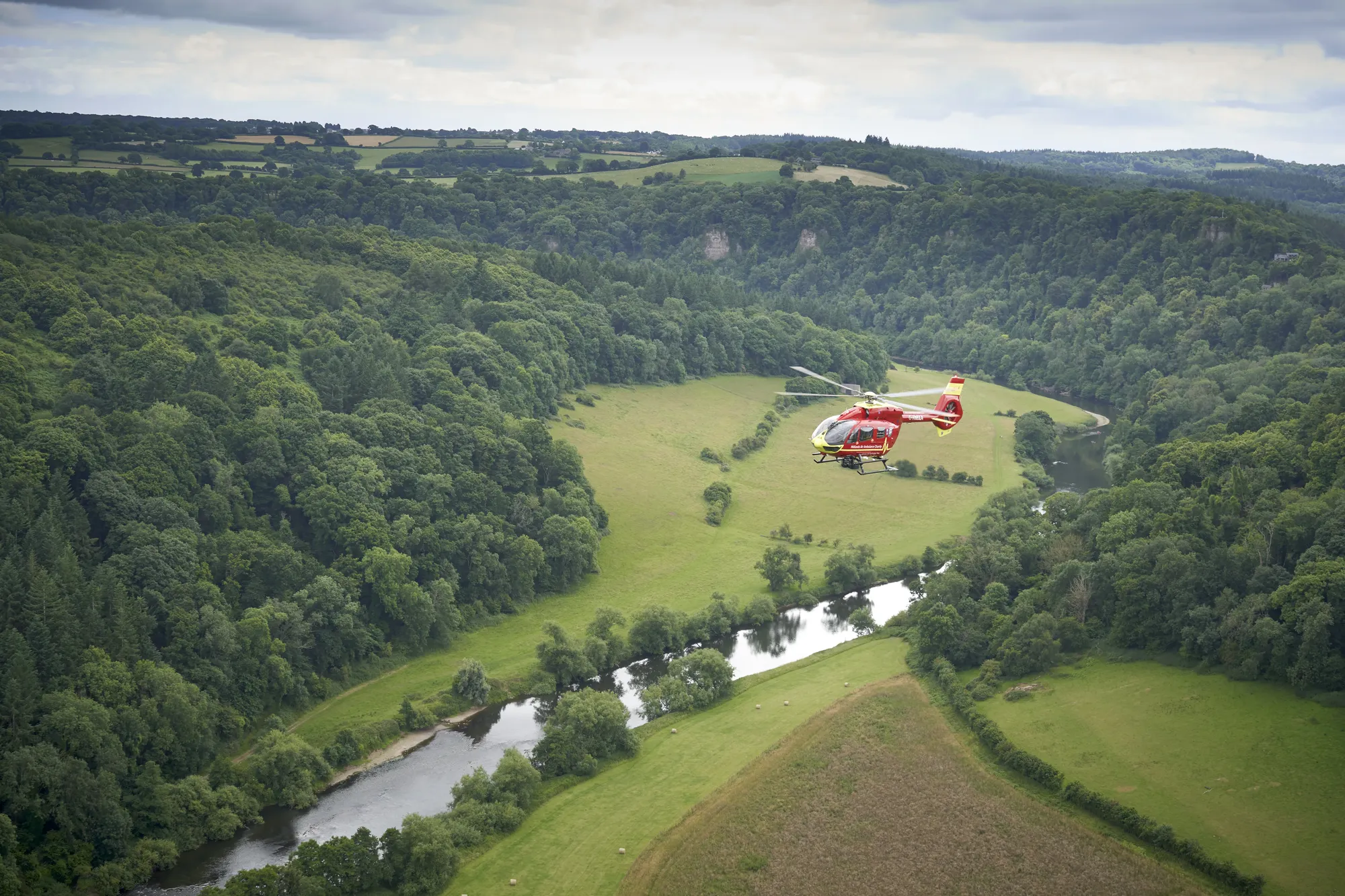 Maac Helicopter Flying Over A Lush Green Valley With A River
