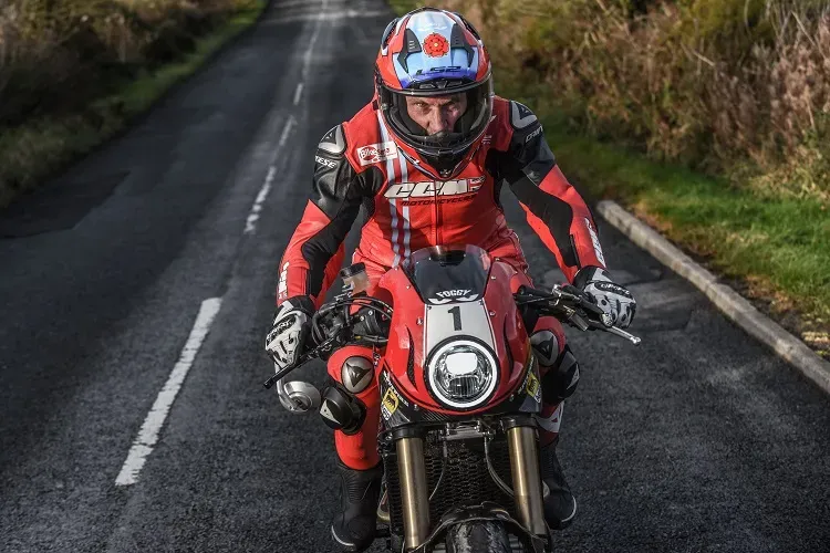 Maac Man In Red And Black Riding A Red Motorcycle Down A Road