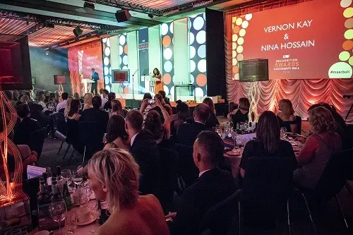 Maac People Sitting At Tables In A Room With A Stage And A Speaker