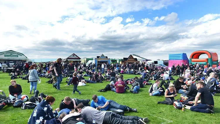 Maac People Sitting On The Grass In A Park With A Large Sign In The Background