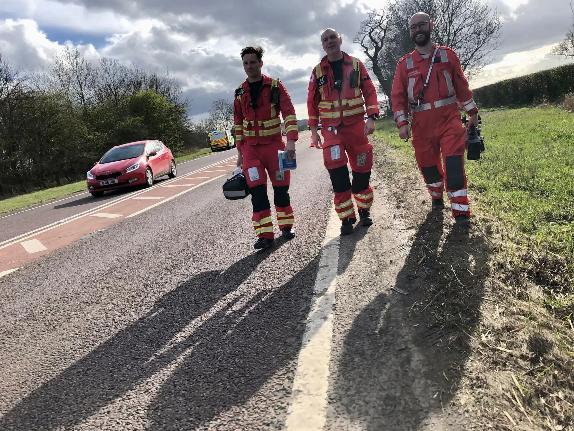 Maac Three Men In Red Uniforms Walking Down A Road With A Car Behind Them