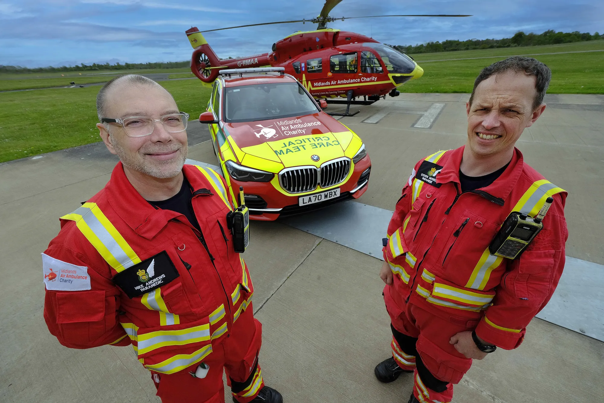 Maac Two Men In Red Uniforms Standing Next To A Yellow Car
