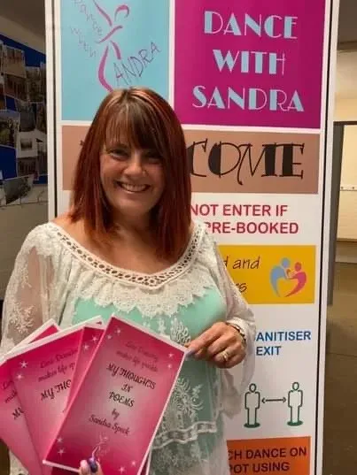Maac Woman Holding Up Three Pink Books In Front Of A Sign