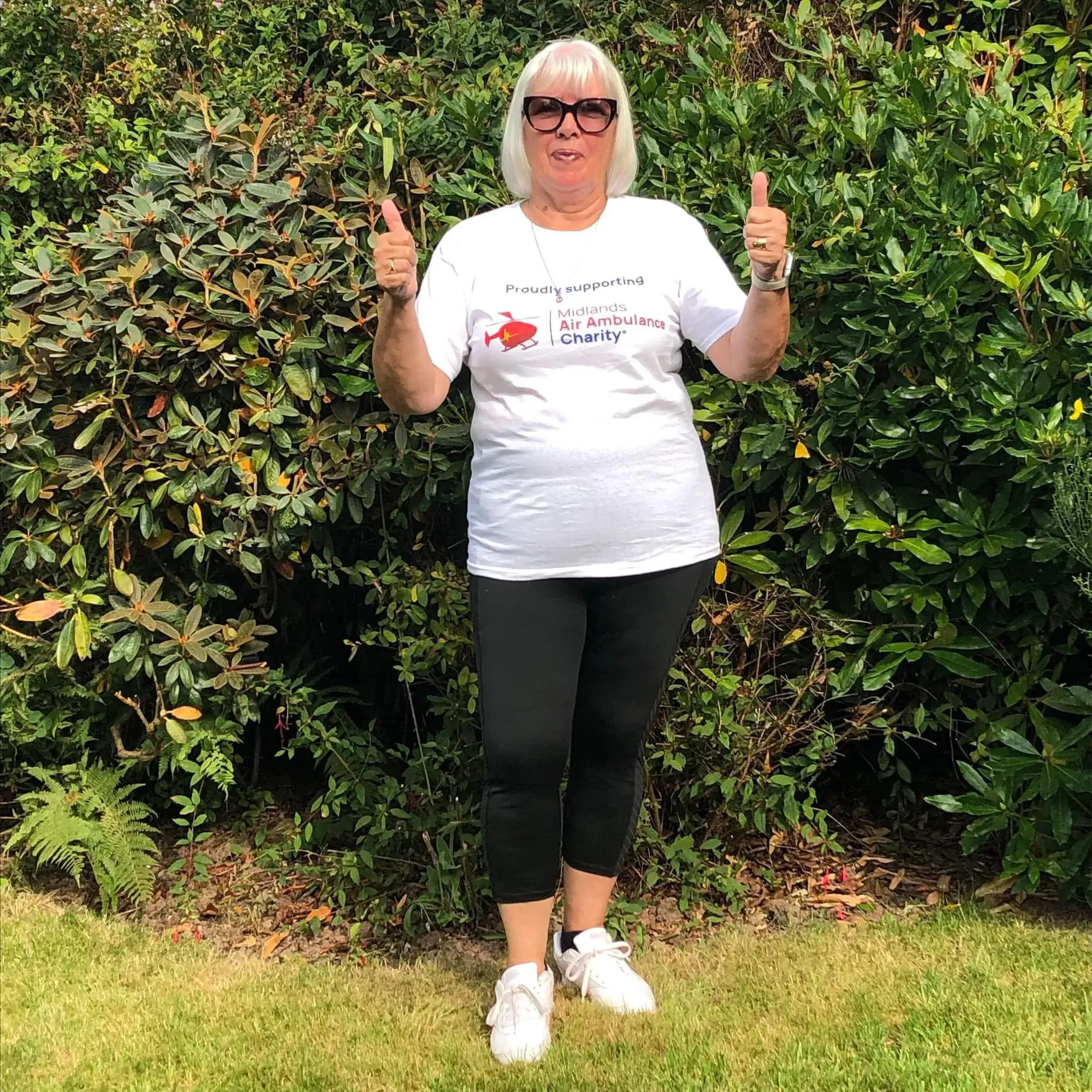 Maac Woman In White Shirt And Black Leggings Standing In Front Of Bushes